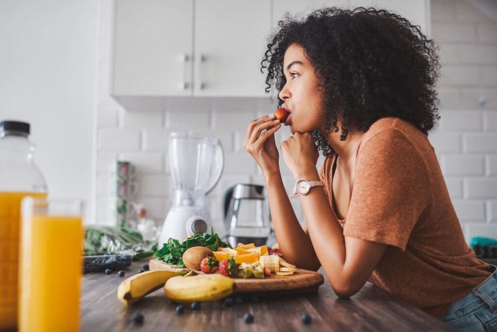 Mulher na cozinha, apoiada em uma balcão, enquanto consome frutas ricas em eletrólitos