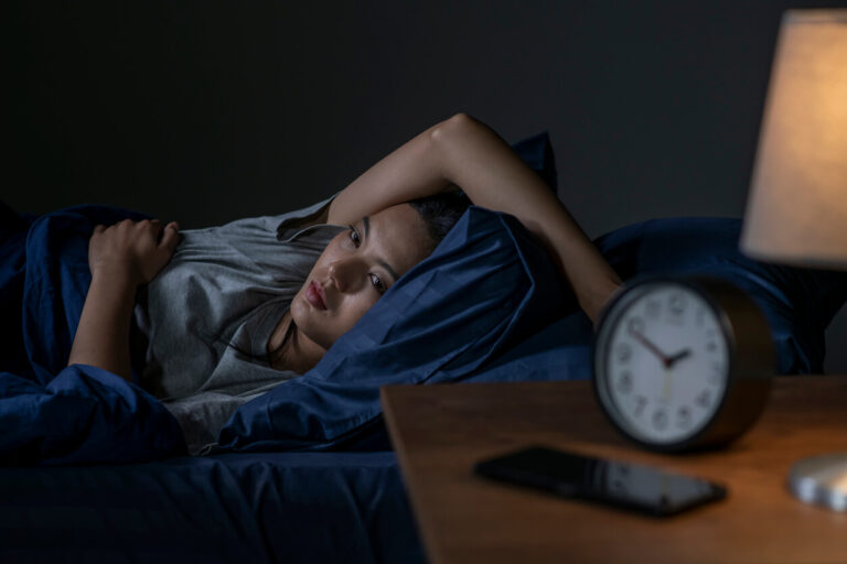 Mulher deitada na cama, com dificuldade para dormir e expressão de cansaço, ilustrando a relação entre intestino e sono.