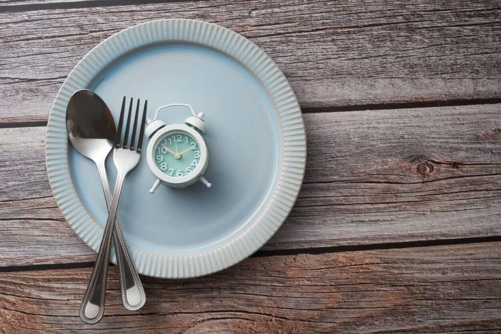 Jejum intermitente e o impacto na microbiota intestinal jejum intermitente Prato com talheres e relógio analógico, simbolizando o conceito de jejum intermitente e controle do tempo das refeições.
