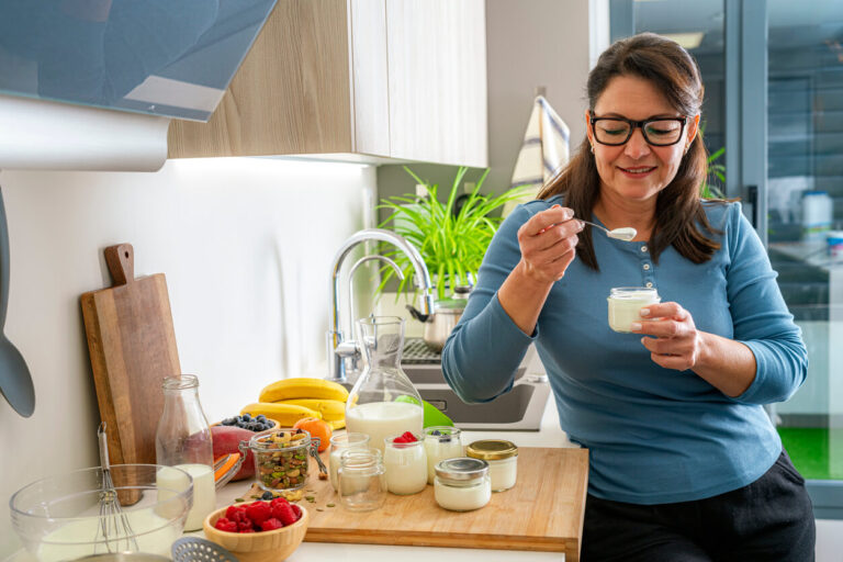 Mulher consumindo iogurte natural em casa, ao lado de frutas e sementes, representando formas de como tomar probiótico no dia a dia e incluí-lo na rotina.
