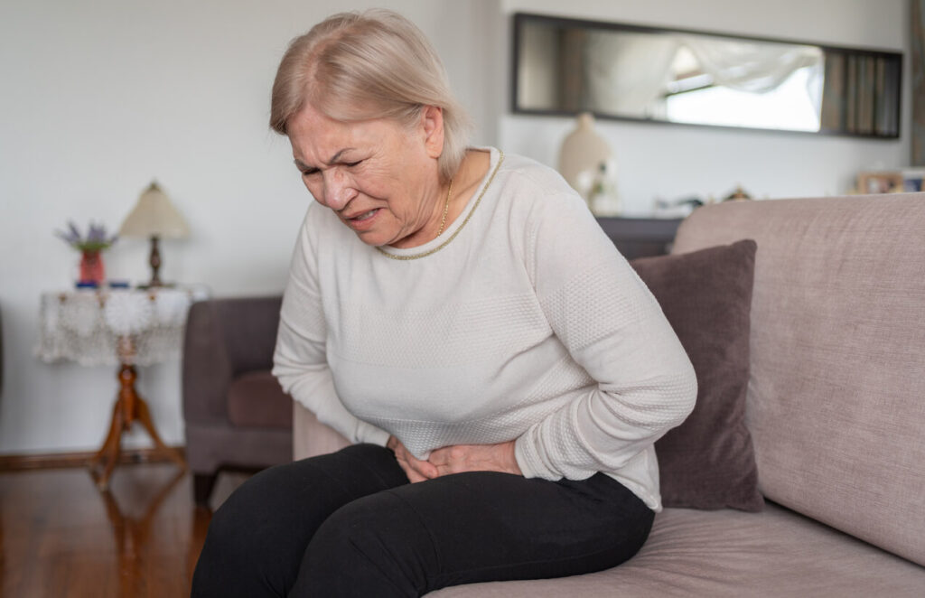 Idosa sentada no sofá com expressão de dor abdominal, ilustrando a importância de cuidar da saúde do intestino na terceira idade para manter qualidade de vida.